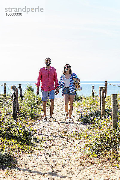 Glückliches Paar mit Sonnenbrille  das sich beim Strandspaziergang an den Händen hält