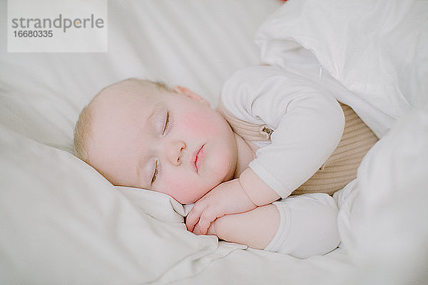 Baby-Mädchen schläft friedlich in weißen Bett mit Blick auf die Kamera mit den Händen für