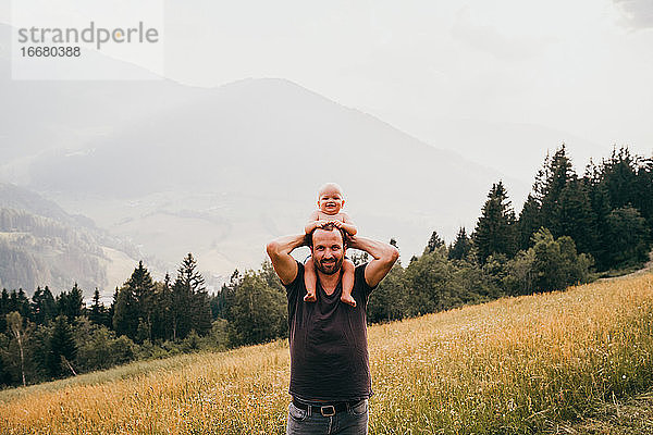 Mann mit Baby auf den Schultern lächelnd mit Blick auf die Berge