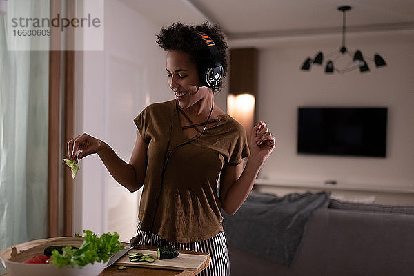 Zufriedene afroamerikanische Frau mit Kopfhörern beim Kochen von Salat