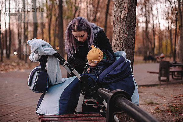 Junge Mutter  die ihren kleinen Sohn im Herbst im Kinderwagen in den Park bringt