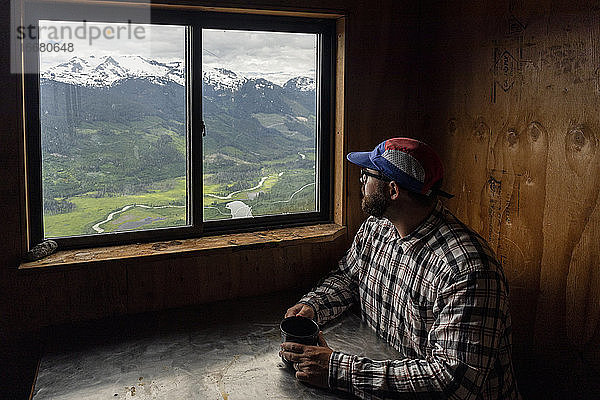 Bärtiger Mann in kariertem Hemd und Mütze sitzt mit einem Becher Heißgetränk am Tisch und blickt auf die Berge  während er sich in einer gemütlichen Hütte in British Columbia  Kanada  ausruht