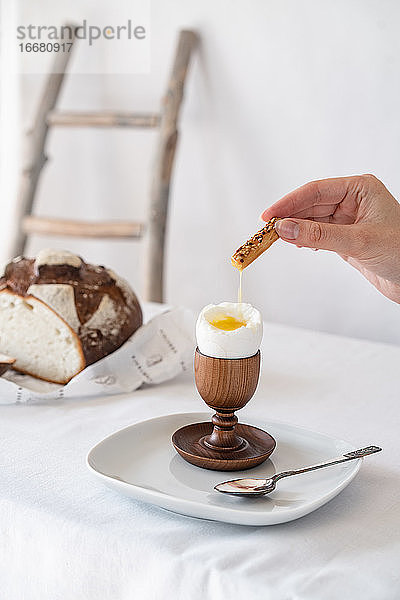 Eine Hand taucht einen Cracker in das Eigelb eines zerbrochenen gekochten Eies auf einem Holzständer auf einem Tisch mit weißem Tischtuch
