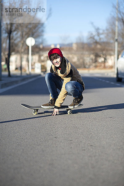 Männlicher Skateboarder  der auf einem Skateboard sitzt und die Kamera im Freien betrachtet