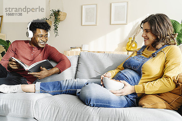 Schwangere Frau isst auf der Couch mit ihrem Mann  der Musik hört und ein Buch liest. Interracial Paar Konzept