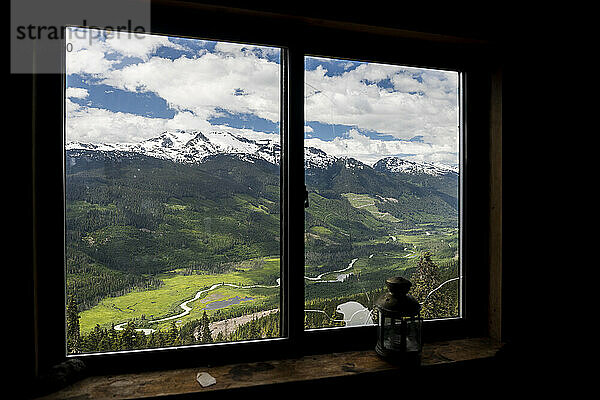 Verschneiter Bergkamm und grünes Tal mit kurvenreicher Straße hinter dem Fenster einer Holzhütte mit Retro-Laterne im Hochland von British Columbia  Kanada