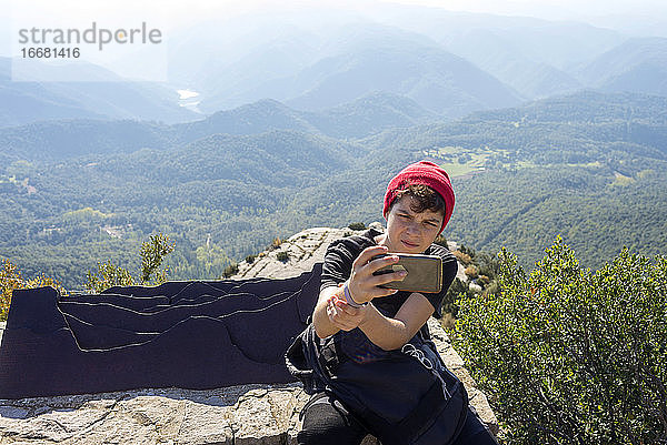 Junger Mann mit roter Wollmütze sitzt am Berg und macht ein Selfie