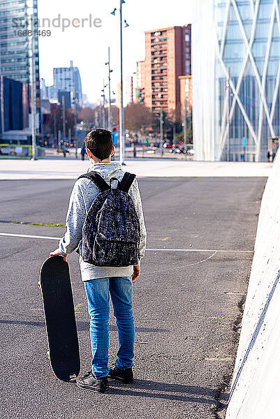 Rückansicht eines Jungen mit Rucksack und Skateboard auf der Straße stehend