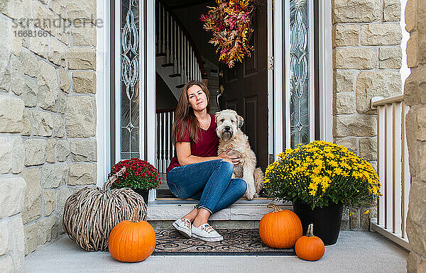 Frau und ihr Hund sitzen in der Tür eines herbstlich dekorierten Hauses.