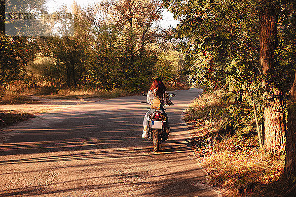 Rückansicht einer Frau auf einem Motorrad auf einer Landstraße inmitten von Bäumen