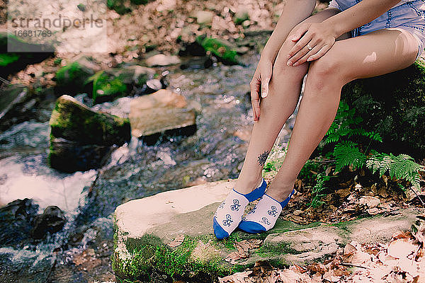 junges Hipster-Mädchen sitzt auf einem Felsen am Fluss im Wald in bedruckten hellen Socken