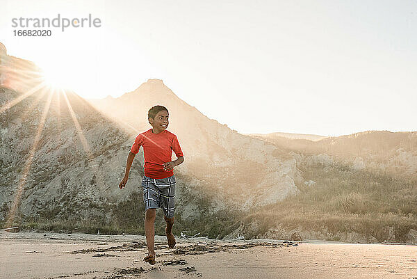 Latino tween Junge läuft am Strand mit Bergen im Hintergrund