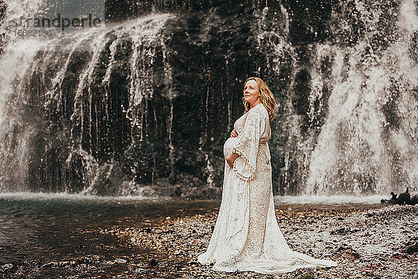 Schwangere Frau steht vor einem Wasserfall im Winter und berührt ihren Bauch