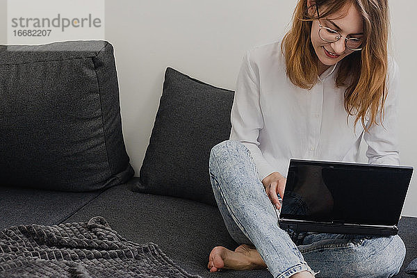 Attraktive junge Frau  die von zu Hause aus arbeitet - Unternehmerin  die auf dem Sofa mit einem Laptop sitzt und ihr Handy von zu Hause aus überprüft