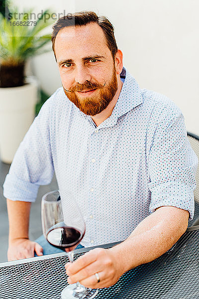 Rothaariger Mann mit Schnurrbart sitzt am Tisch mit einem Glas Rotwein