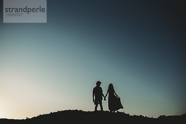 Silhouette von Bruder und Schwester  die auf einer Sanddüne stehen und sich an den Händen halten