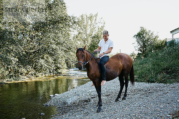 Porträt eines jungen blonden Mannes  der auf einem Pferd über einen Fluss reitet