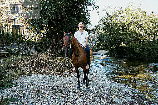 Porträt eines jungen blonden Mannes  der auf einem Pferd über einen Fluss reitet