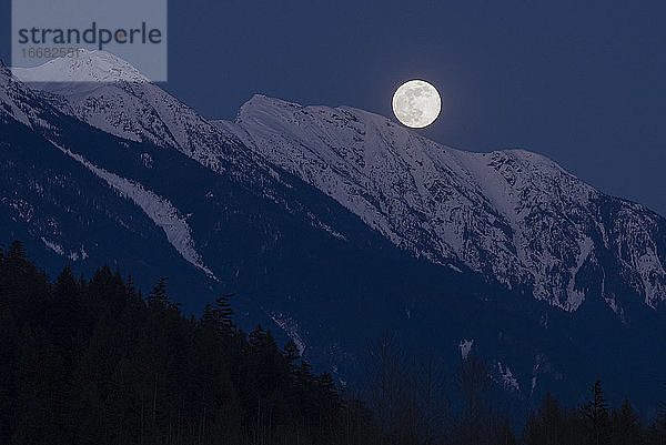 Der Vollmond geht in einer Frühlingsnacht in den Coast Mountains von British Columbia über schneebedeckten Berggipfeln auf.