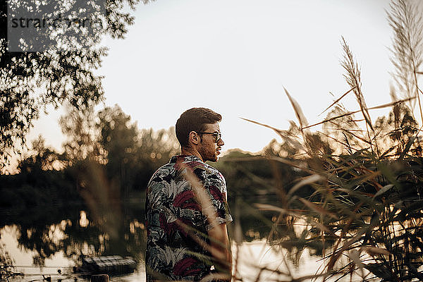 Junger Mann mit Sonnenbrille auf dem Rücken an einem See bei Sonnenuntergang