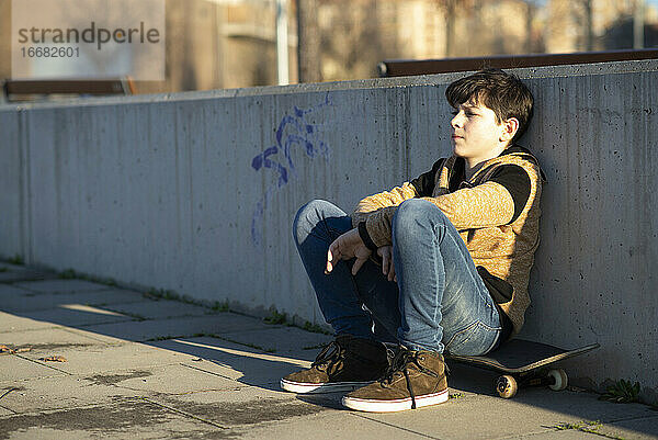 Junger Teenager sitzt auf seinem Skateboard und lehnt sich an einen Zaun