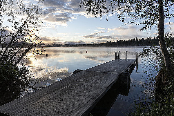Dock von ruhigen See und Baum bei Sonnenaufgang mit Reflexion der Wolken