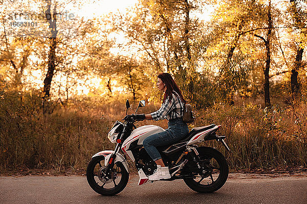 Junge selbstbewusste Frau fährt Motorrad auf Landstraße bei Sonnenuntergang