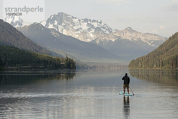 Rückenansicht eines anonymen Mannes auf einem Paddelbrett auf der ruhigen Oberfläche des Duffey-Sees und Genießen der Berglandschaft am Morgen in British Columbia