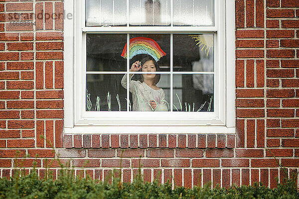 Blick von außen auf ein kleines Kind  das im Fenster steht und einen Regenbogen malt