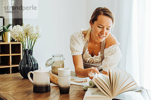 Frau liest ein Buch mit Rezepten  während sie am Küchentisch kocht