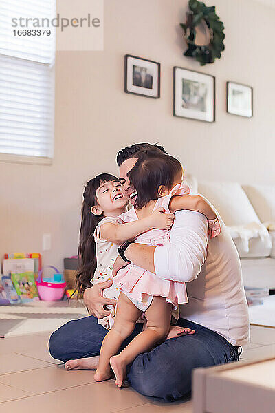 Der Vater sitzt auf dem Boden und umarmt seine beiden Töchter.