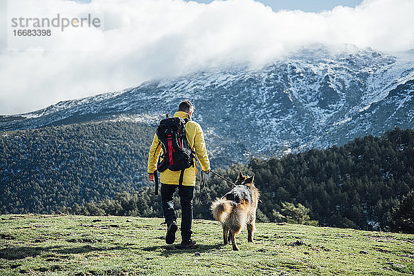 Junger Mann mit gelber Jacke und Rucksack spielt mit deutschem Schäferhund in den Bergen.