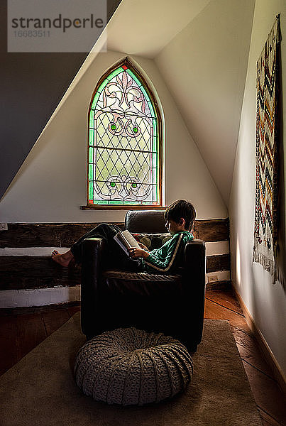 Junger Junge liest in einem Ledersessel vor einem verschnörkelten Fenster eines Hauses.