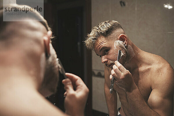 Ein blonder Mann rasiert sich mit einem Rasiermesser im Badezimmer