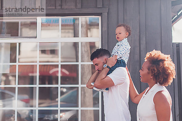 Papa trägt Baby auf den Schultern mit Mama lächelnd vor großem Fenster