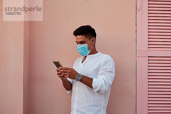 Junger lateinamerikanischer Mann mit Maske schaut auf sein Telefon auf rosa Hintergrund