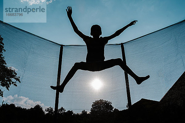 Horizontales Gegenlichtfoto einer Silhouette eines barfüßigen Jungen  der auf einem Trampolin mit Netz springt oder fliegt  wobei die Sonne im Hintergrund die Sonnenstrahlen auf seinem Schatten reflektiert