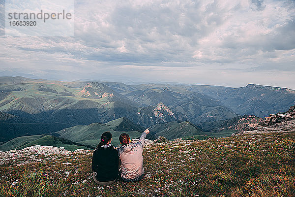 Zwei Männer sitzen und schauen in die Ferne und auf die Berge  diskutieren etwas und zeigen mit der Hand irgendwo in die Ferne.