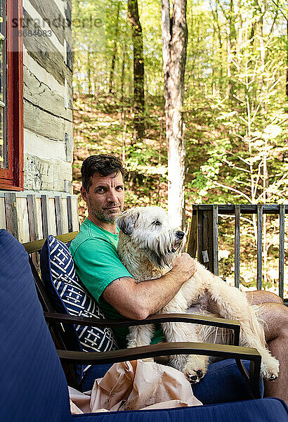 Mann  der einen flauschigen Hund auf seinem Schoß hält  draußen auf einer Terrasse im Wald.