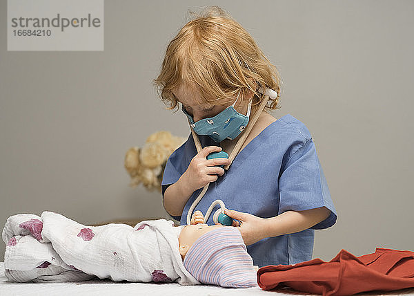 Ein kleines Kind  das eine medizinische PSA trägt  untersucht eine Babypuppe  indem es mit einem Stethoskop ihre Vitalfunktionen prüft.