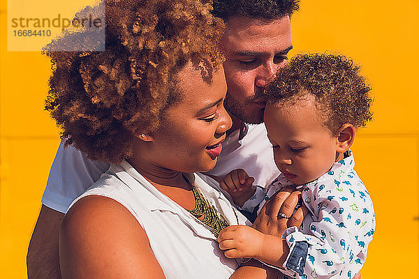 Mama hält Kleinkind und Papa küsst seinen Kopf vor einer gelben Wand