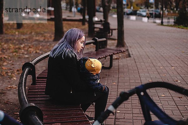 Junge Mutter hält ihr Baby im Arm  während sie auf einer Bank im Park sitzt