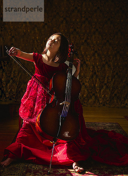 Ein kleines hübsches Kind in einem langen roten Kleid spielt Cello im Fensterlicht