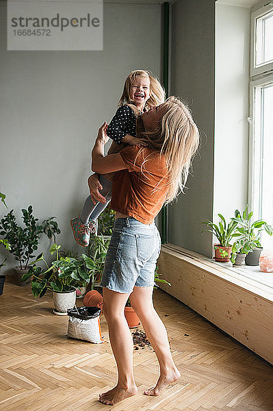 Frau hält in Umarmungen ihr lachendes Kind im Wohnzimmer mit vielen Pflanzen