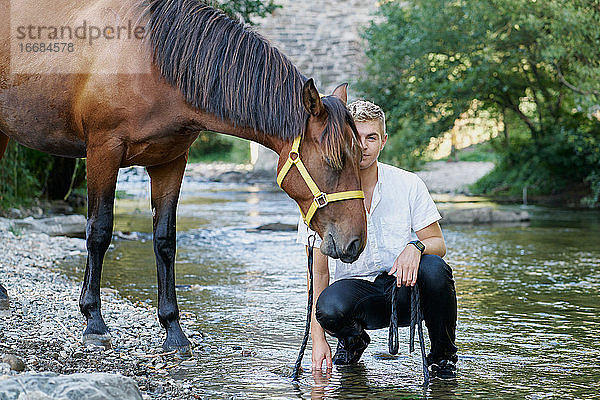 Porträt eines blonden jungen Mannes mit einem Pferd an einem Fluss