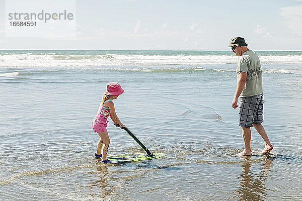 Junges Mädchen und ihr Vater spielen am Strand mit einem Skimboard