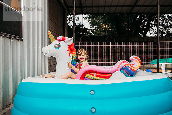 Junges Mädchen sitzt auf einem aufblasbaren Regenbogeneinhorn im Hinterhofpool