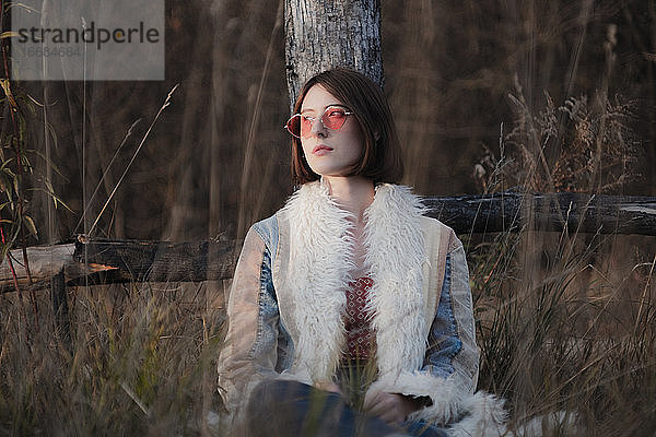 Junge Frau in Hippie-Mantel und roter Sonnenbrille zwischen Unkraut und Gras