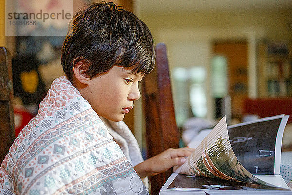 Junge im Profil sitzt in eine Decke gehüllt am Tisch und betrachtet ein Fotobuch