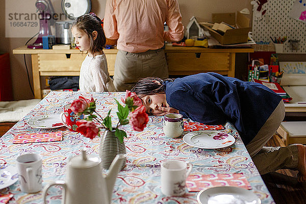Ein Junge im Anzug liegt über einem für den Tee gedeckten Tisch  im Hintergrund die Familie